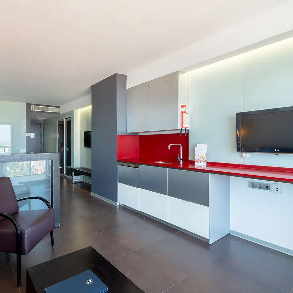 Zona de estar de la Suite Prestige con televisión de pared, kitchenette roja y sillón de cuero.